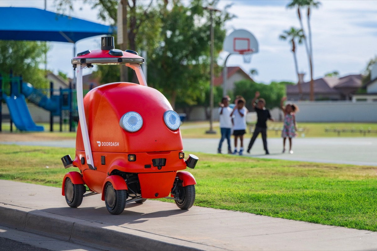 A red DoorDash sidewalk delivery robot parked at a curb near a playground.